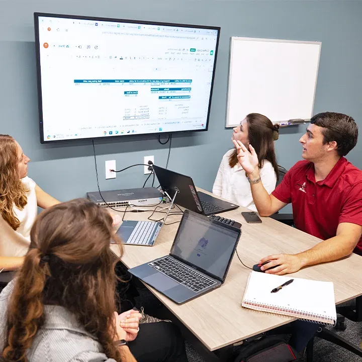 Four students gathered at table with laptops and notebooks looking at a spreadsheet on TV screen.